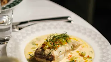 A plated gourmet dish featuring fish in a creamy sauce topped with herbs, served on a white tablecloth alongside a bowl of roasted vegetables and a glass of water.