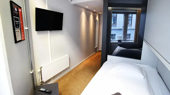 Valoisa ja kapea hotellihuone, jossa on yhden hengen vuode, seinälle asennettu tv ja ikkuna taustalla.