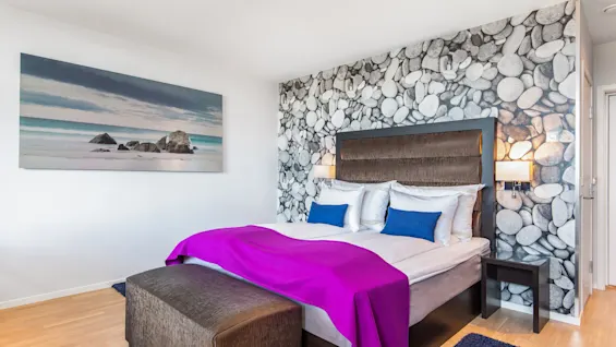En dobbeltseng med lilla teppe og blå puter står mot en vegg med steinmønster, ved siden av et strandlandskapsmaleri, i et lyst, moderne rom.