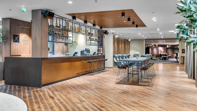 A modern hotel lobby with wooden floors, a coffee bar counter, stools, and tables; warm lighting creates a welcoming atmosphere.