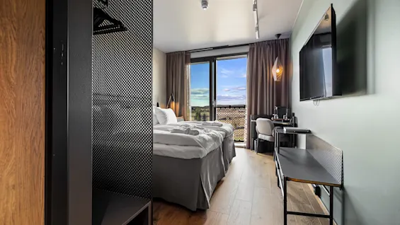 Ett prydligt hotellrum har en dubbelsäng som vetter mot en väggmonterad TV. Dagsljus strömmar in genom ett fönster med gardiner, vilket skapar en inbjudande atmosfär.