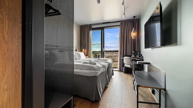 Et pent ordnet hotellrom har en dobbeltseng vendt mot en veggmontert TV. Dagslys kommer inn gjennom et vindu med gardiner, og skaper en innbydende atmosfære.