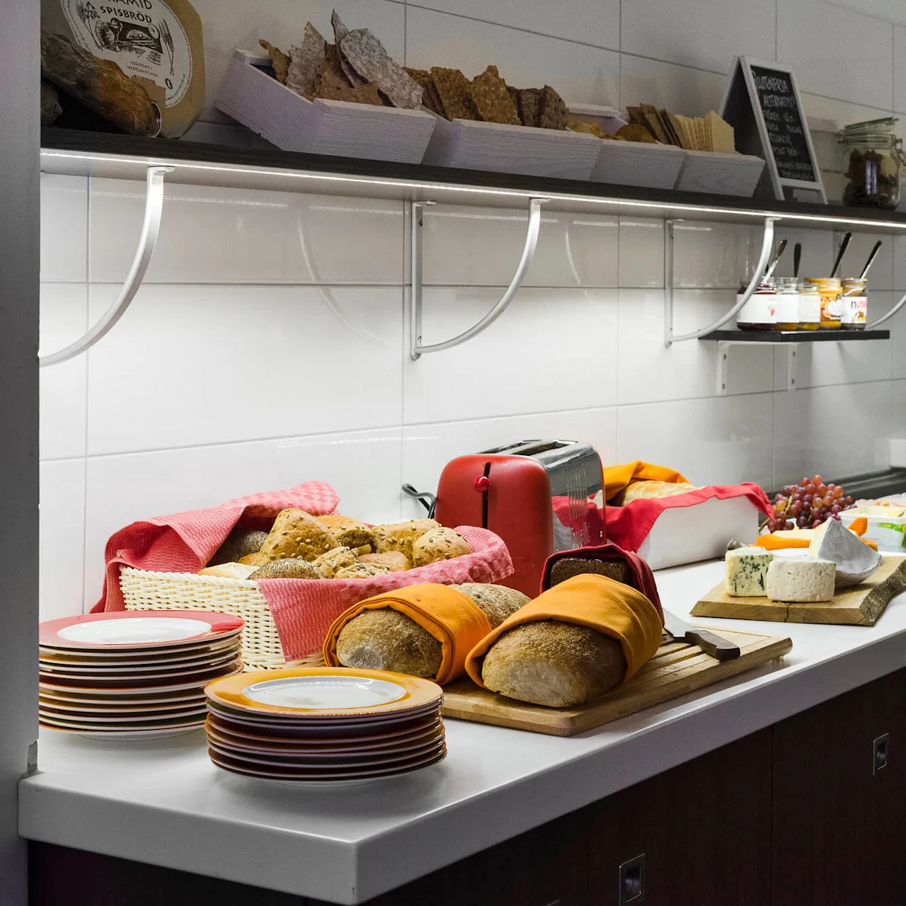 En frokostbuffé viser frem ulike brød, oster, frukter og kjøtt i et rent rom med hvite fliser.