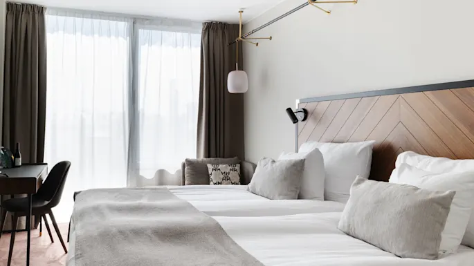 A neatly made bed with a grey blanket and multiple pillows against a wooden headboard, with sheer curtains on a window and a table with a chair.