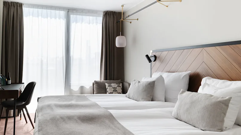 A neatly made bed with a grey blanket and multiple pillows against a wooden headboard, with sheer curtains on a window and a table with a chair.