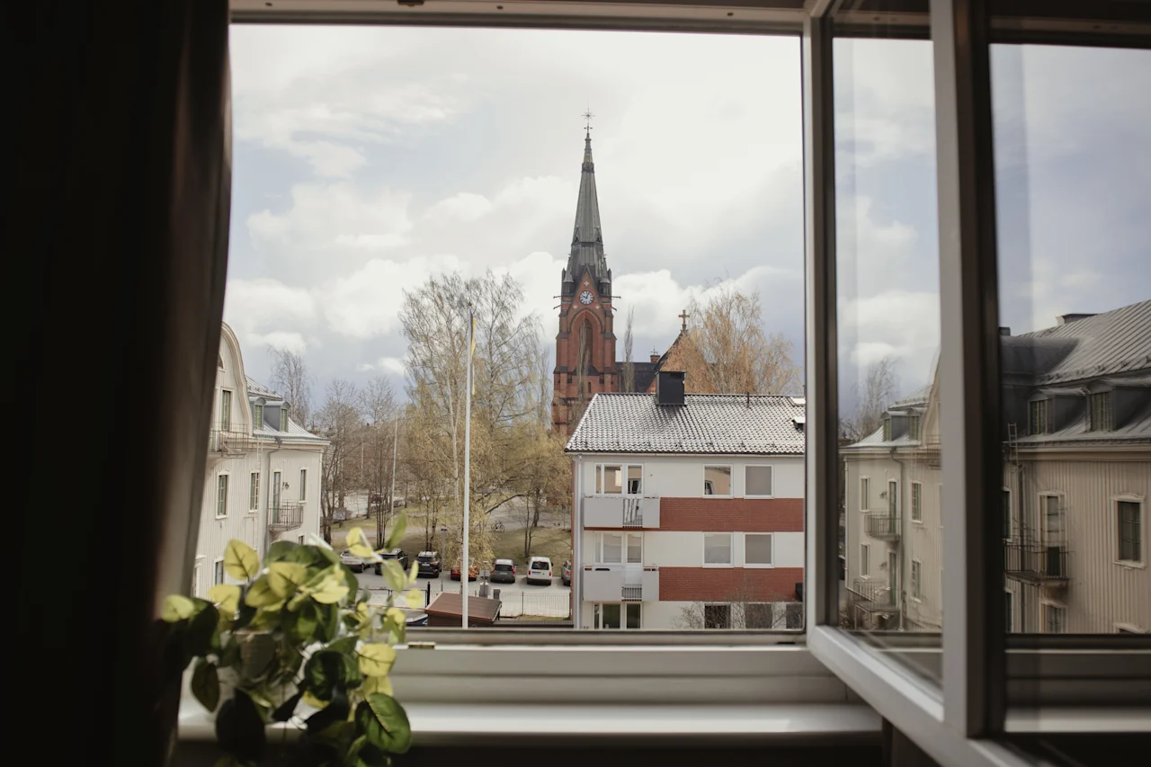 A church tower stands tall outside an open window, surrounded by trees and residential buildings. A potted plant rests on the windowsill.