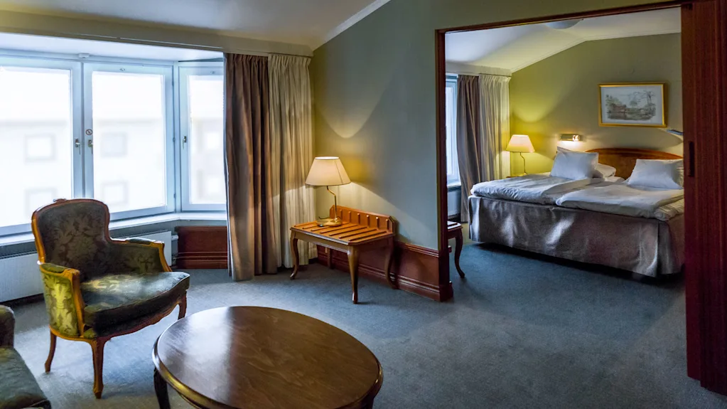 Ett mysigt hotellrum har en stor säng med vita lakan, sänglampor och en sittgrupp med fåtöljer och ett soffbord vid ett fönster.
