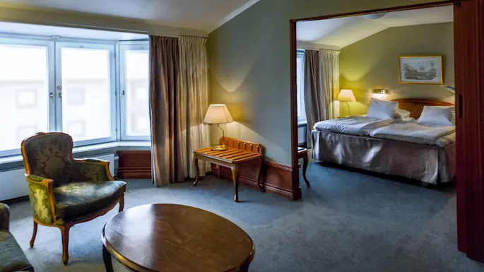 Et koselig hotellrom har en stor seng med hvitt sengetøy, nattbordslamper og en sittegruppe med lenestoler og et kaffebord ved et vindu.