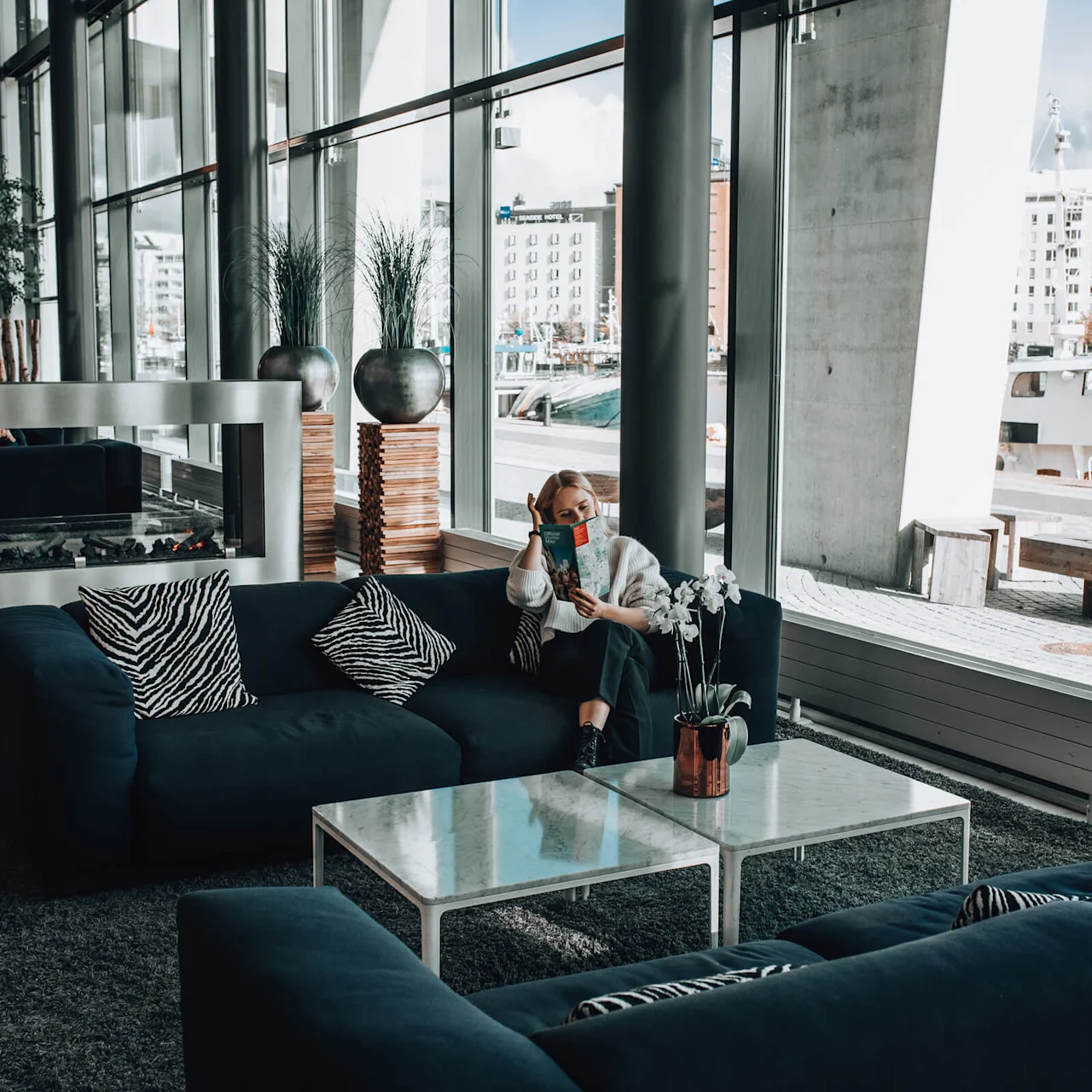 En person læser et magasin på en sofa i en moderne lounge med store vinduer, zebramønstrede puder og udsigt over en vandfront udenfor.