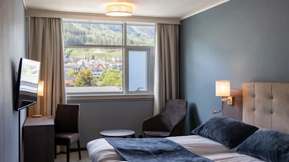 Ett hotellrum med dubbelsäng, marina kuddar och filt, ligger bredvid ett fönster med utsikt över en bergsstad. En stol och ett skrivbord finns till vänster.