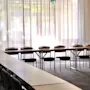 Long tables are arranged with chairs underneath in an empty conference room. Sunlight filters through sheer curtains, illuminating the carpeted floor.
