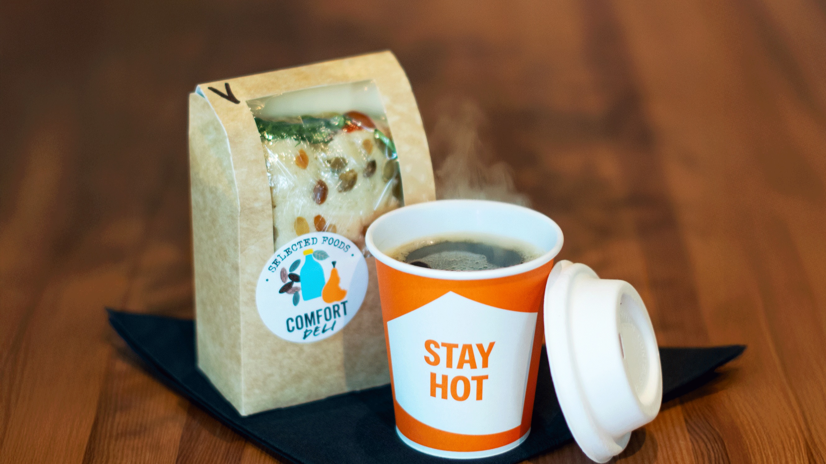 A steaming cup of coffee with 'STAY HOT' on it, next to a packaged sandwich labeled 'SELECTED FOODS DELI' on a wooden table.