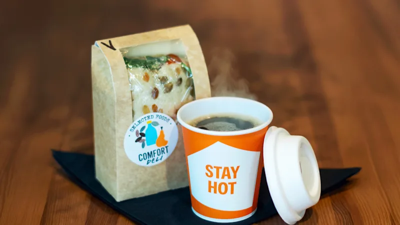 En dampende kop kaffe med 'STAY HOT' på, ved siden af en pakket sandwich mærket 'SELECTED FOODS DELI' på et træbord.