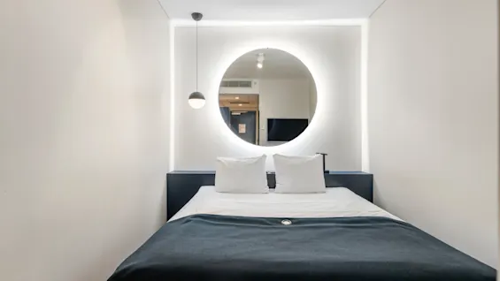Et moderne hotelværelse med en seng, hvide puder og en mørkeblå dyne. Et rundt spejl med glødende lys hænger over sengen.