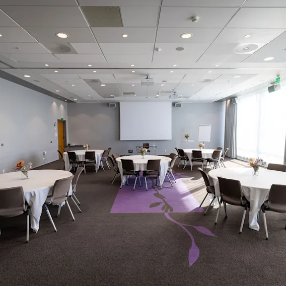 Runde bord med hvite duker og stoler er plassert i et konferanserom. En projektorskjerm er montert foran, med store vinduer på høyre side.