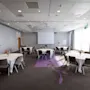 Pyöreät pöydät valkoisilla liinoilla ja tuoleilla on järjestetty kokoushuoneessa. Projektorin näyttö on asennettu eteen, suuret ikkunat ovat oikealla puolella.