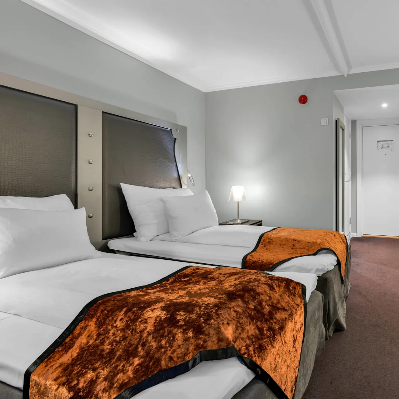 To pænt redte enkeltsenge med orange tæpper står ved grå vægge. Et skrivebord med en gennemsigtig stol og lampe står i hjørnet og skaber en ryddelig hotelfølelse.
