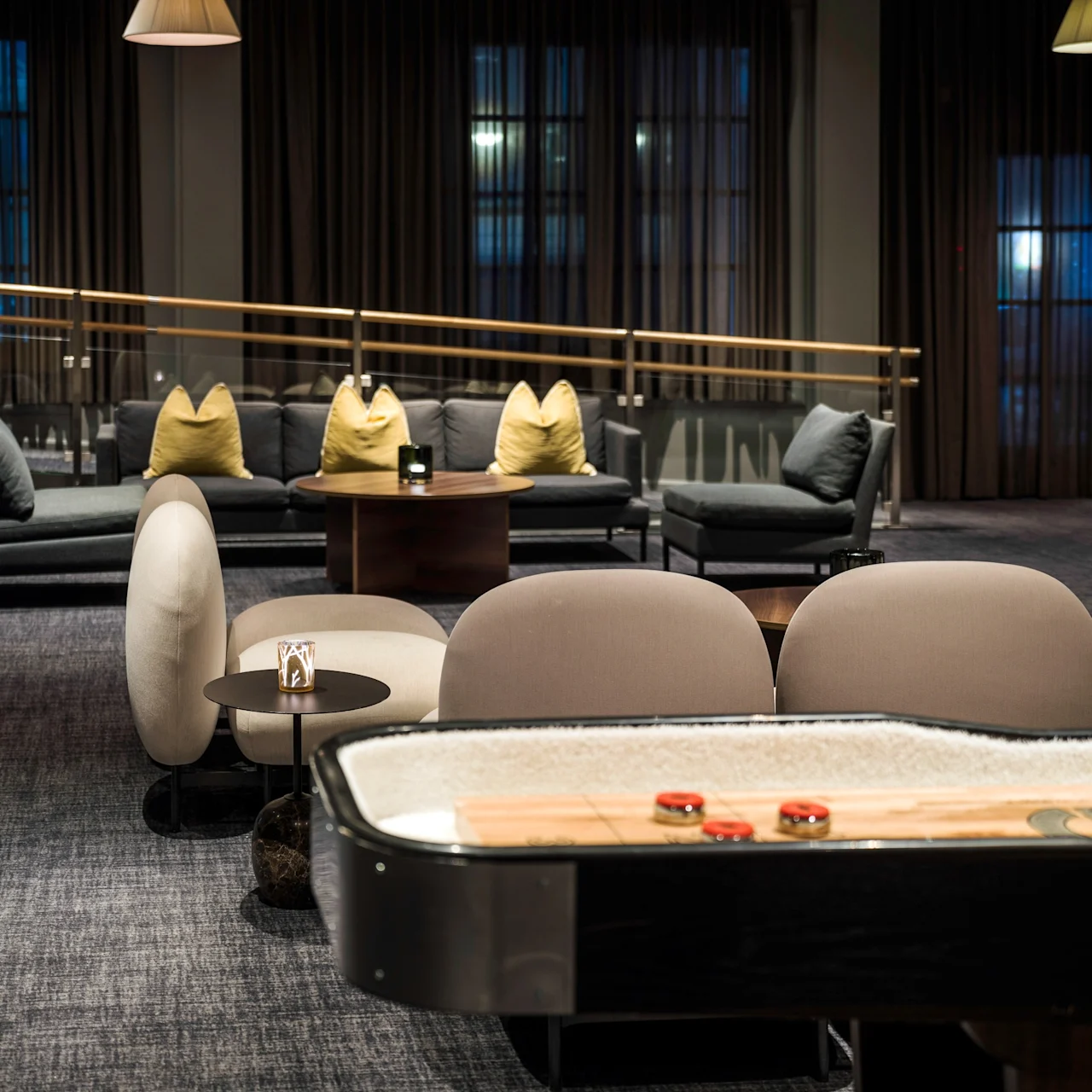 Shuffleboard table, with red pucks, stands in a dimly lit lounge area. Surrounding are modern chairs and a sofa with yellow pillows. Dark curtains and soft lighting create a cozy ambiance.