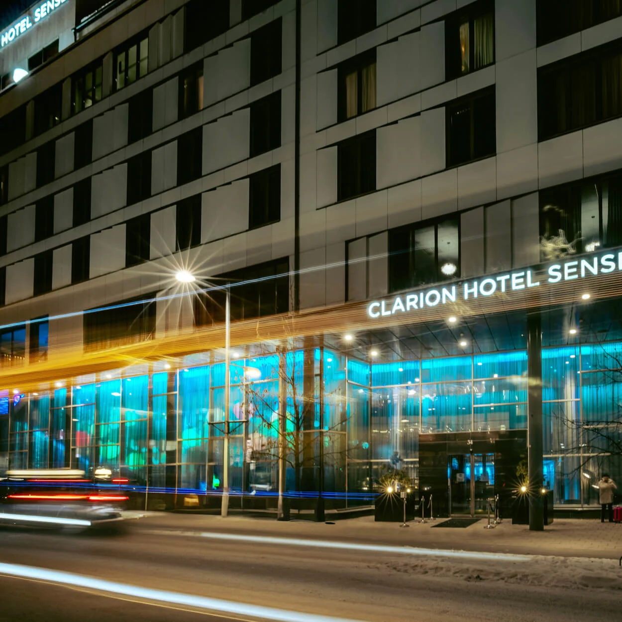 Fasaden på ett upplyst hotell med skylten "CLARION HOTEL SENSE". Bilar med ljusspår kör förbi under en snöig natt. En röd Clarion Hotel-skylt syns.