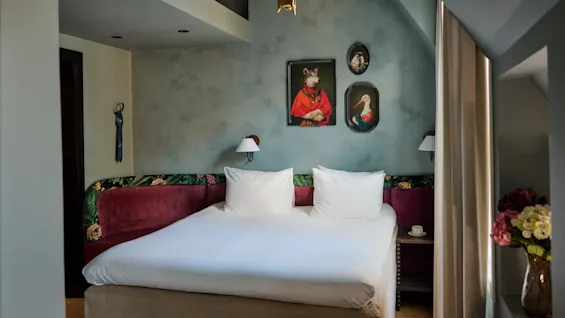 A bed with white linens stands against a gray wall adorned with quirky animal portraits. It is flanked by bedside tables and a deep red couch, with curtains drawn aside.