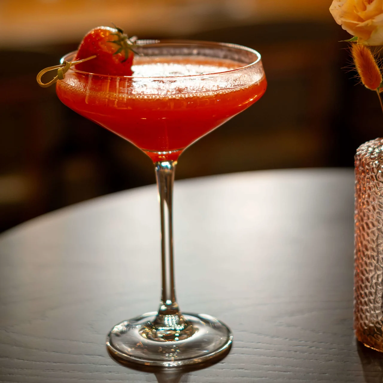 A cocktail glass holds a red drink garnished with a fresh strawberry on a skewer, set on a wooden table beside a vase with pale yellow flowers in a softly lit setting.