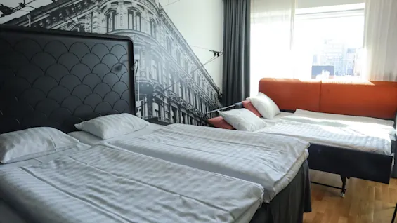 Två prydligt bäddade sängar står i ett modernt hotellrum med en stor väggmålning av en gammal byggnad. Ett fönster ger stadsutsikt och lyser upp den minimalistiska inredningen.