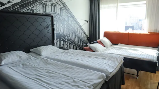 Två prydligt bäddade sängar står i ett modernt hotellrum med en stor väggmålning av en gammal byggnad. Ett fönster ger stadsutsikt och lyser upp den minimalistiska inredningen.