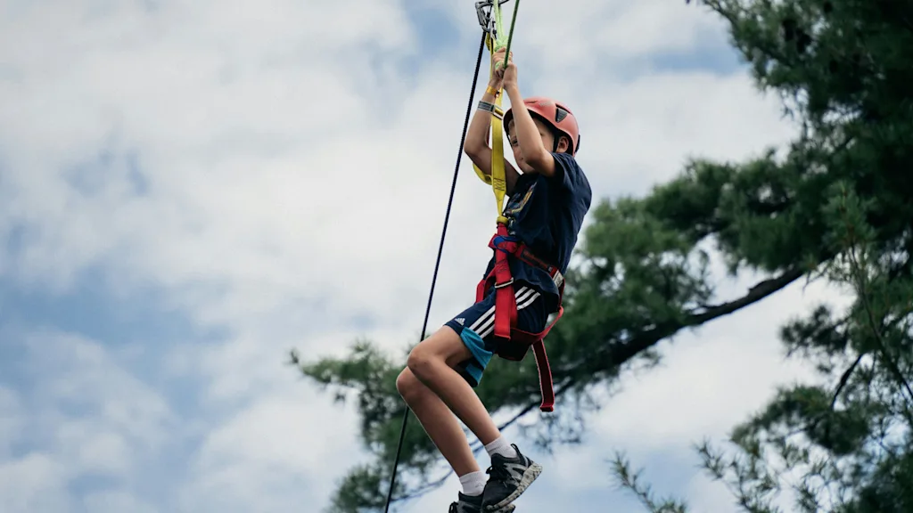 A person in a helmet and harness is actively engaged in zip-lining, soaring through the air with trees and a cloudy sky visible.