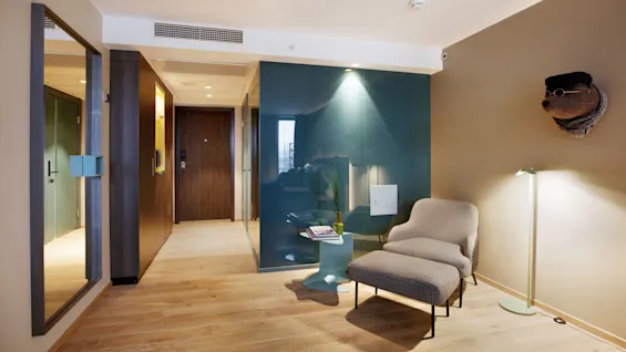 A lounge chair with footstool sits under a wall-mounted bear sculpture wearing glasses. A small side table holds magazines near a floor lamp on wooden flooring.