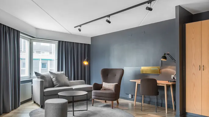 En grå sofa med puter er plassert ved et stort vindu med gardiner. I nærheten gir en matchende lenestol, rundt kaffebord og skrivebord en koselig stuestemning.