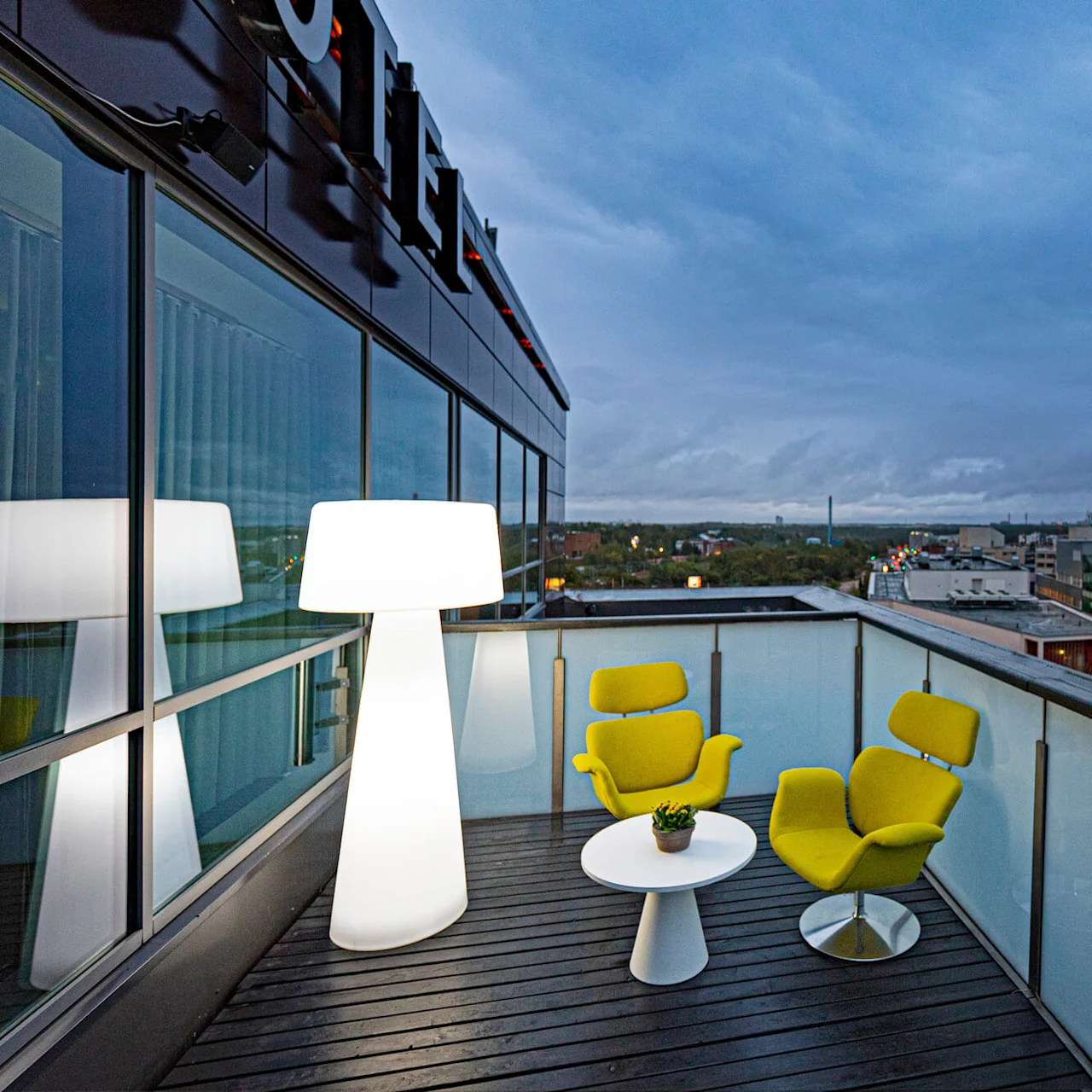 A rooftop terrace features two yellow chairs and a white table with a plant, illuminated by a tall modern lamp, surrounded by glass panels overlooking the city skyline.