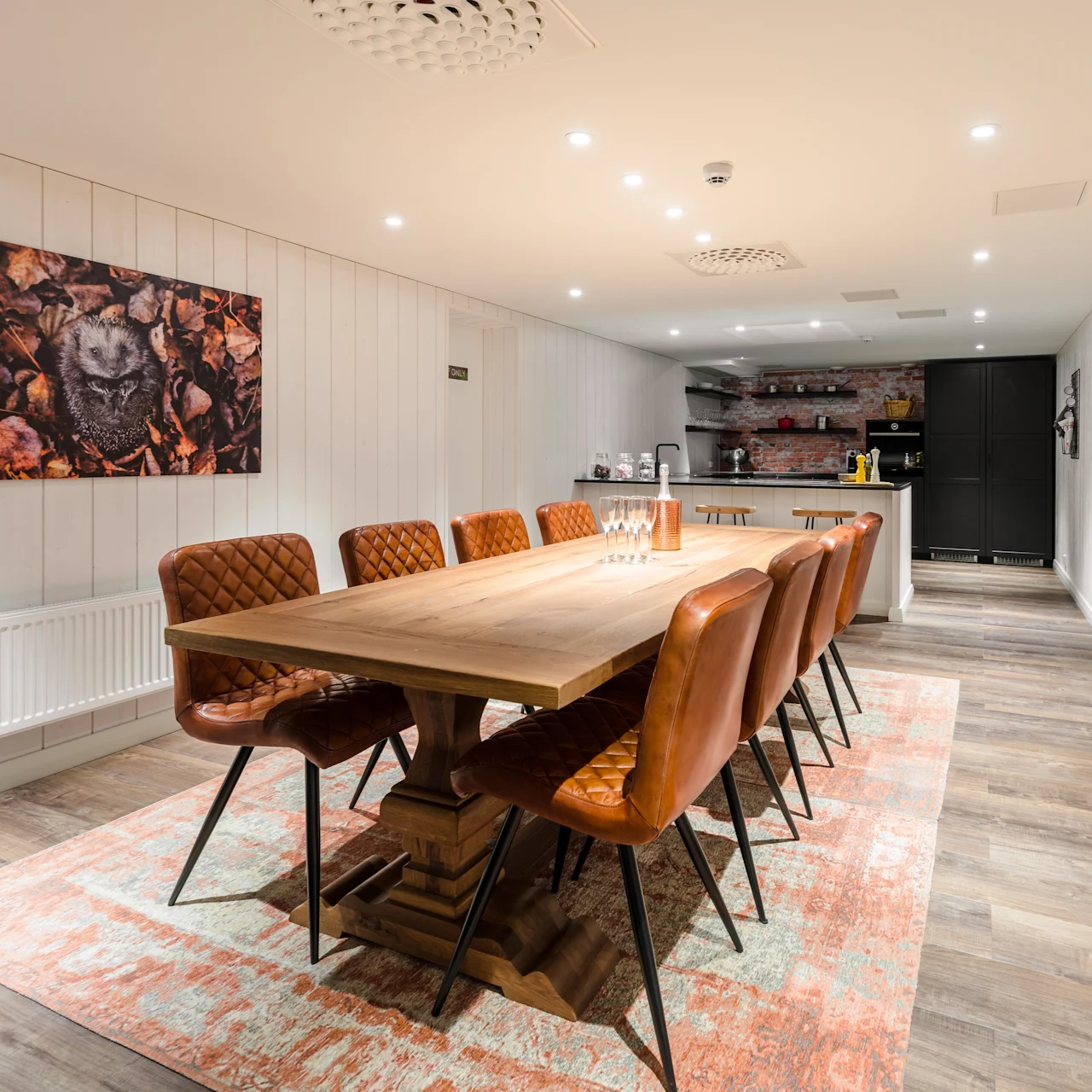 A long wooden dining table with brown leather chairs sits on a patterned rug in a well-lit room. A colorful abstract painting hangs on the white-panelled wall.