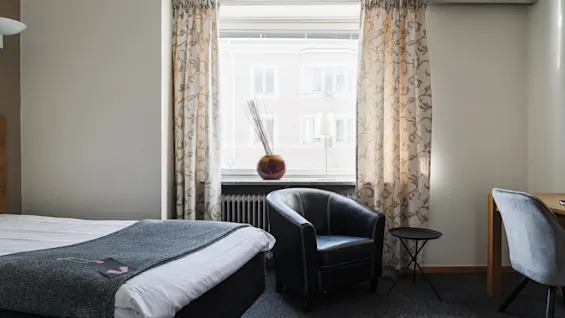 Ett prydligt hotellrum med en säng med grå filt, en svart fåtölj och ett bord bredvid. Ett fönster med mönstrade gardiner lyser upp rummet.