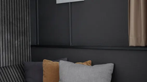 A wall sconce with an angled white shade is mounted above a striped cushioned bench adorned with three colored pillows. Dark walls and a curtain create a cozy interior setting.