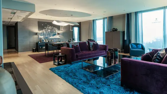A luxurious living room features a purple sofa and chairs situated on a blue rug, with modern decor and large windows. Text on windows: "Clarion Hotel Post" and "Clarion Post".