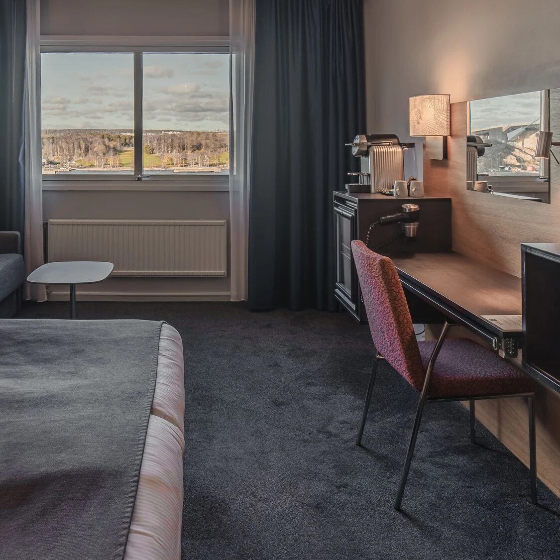 A neatly arranged hotel room features a bed with white linens, a gray sofa with decorative pillows, a desk with a chair, and a large window overlooking a scenic landscape.