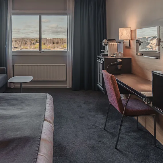 Ett prydligt hotellrum med en säng med vita lakan, en grå soffa med dekorativa kuddar, ett skrivbord med stol och ett stort fönster med utsikt över ett naturskönt landskap.