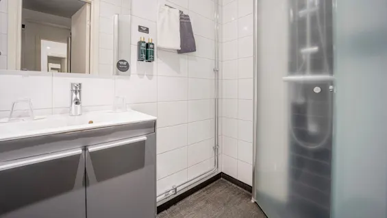 Et badeværelse med håndvask, spejlskab og frostet glasbruser. Håndklæder hænger på en holder over toiletartikler. Væggene er flisebelagte, gulvet er mørkt. Minimalistisk design.