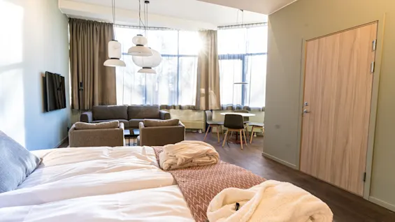 A neatly arranged hotel room features a bed with robes, a sitting area with armchairs and a sofa, a small dining table, and large windows allowing natural light.