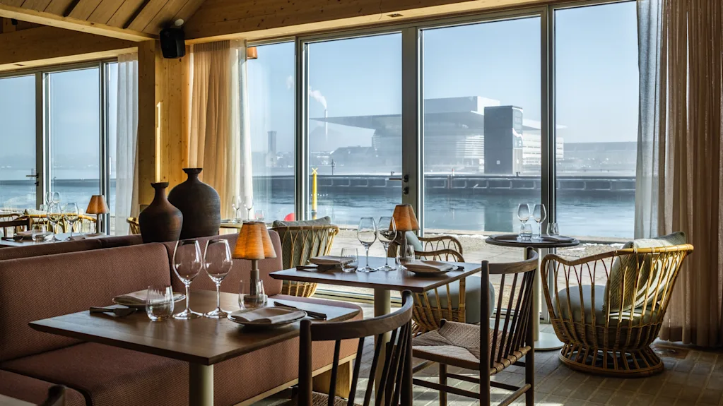 Matsalsbord är dukade med glas och servetter i en mysig restaurang med träinredning. Stora fönster avslöjar en vattensikt med en modern byggnad på en solig dag.