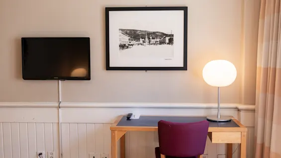 Fladskærms-TV monteret på en beige væg ved siden af indrammet sort-hvid kunst over et træskrivebord med en lilla stol; rund lampe tændt på skrivebordet.