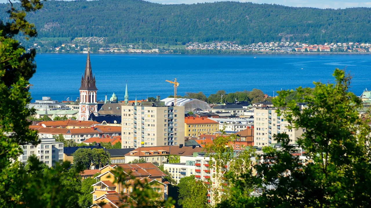 City buildings, including a church with a tall spire, rise among greenery. The landscape opens to a vast blue lake, bordered by forested hills under a clear sky.