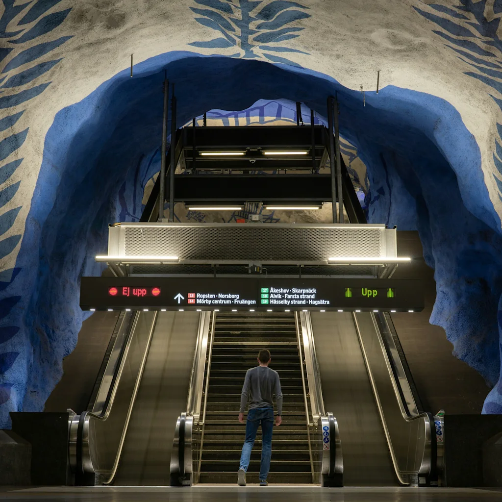 En person står vid rulltrappa och trappor i en tunnelbanestation med blå och vit stockholm subway art. Skyltar visar Ej Upp och Upp.