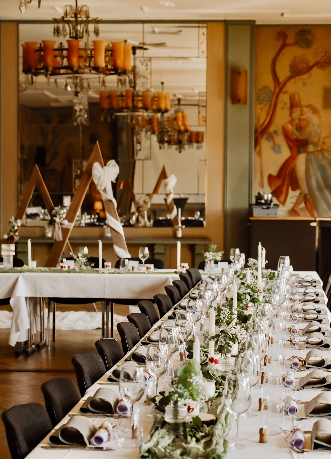 A long, elegantly set dining table with white tablecloths is decorated with green foliage, candles, and glassware. A mural and large chandeliers create a refined dining atmosphere.