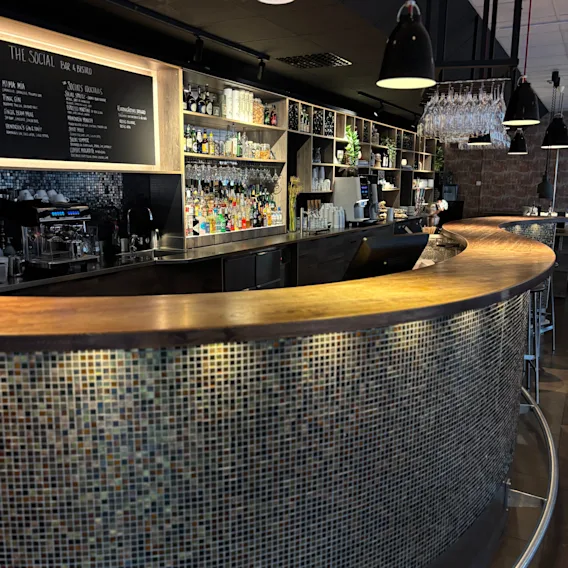 Modern bar med böjd trädisk, kaklad front och flaskhyllor. Menyn: THE SOCIAL BAR & BISTRO SOCIALS COCKTAILS.