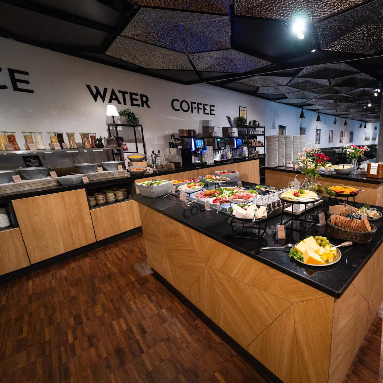 Buffetbord med en række forskellige retter, omgivet af et moderne spiseområde. På væggen står "WATER COFFEE", med korn og drikkevarer på sideborde. Moderne design og dæmpet belysning.