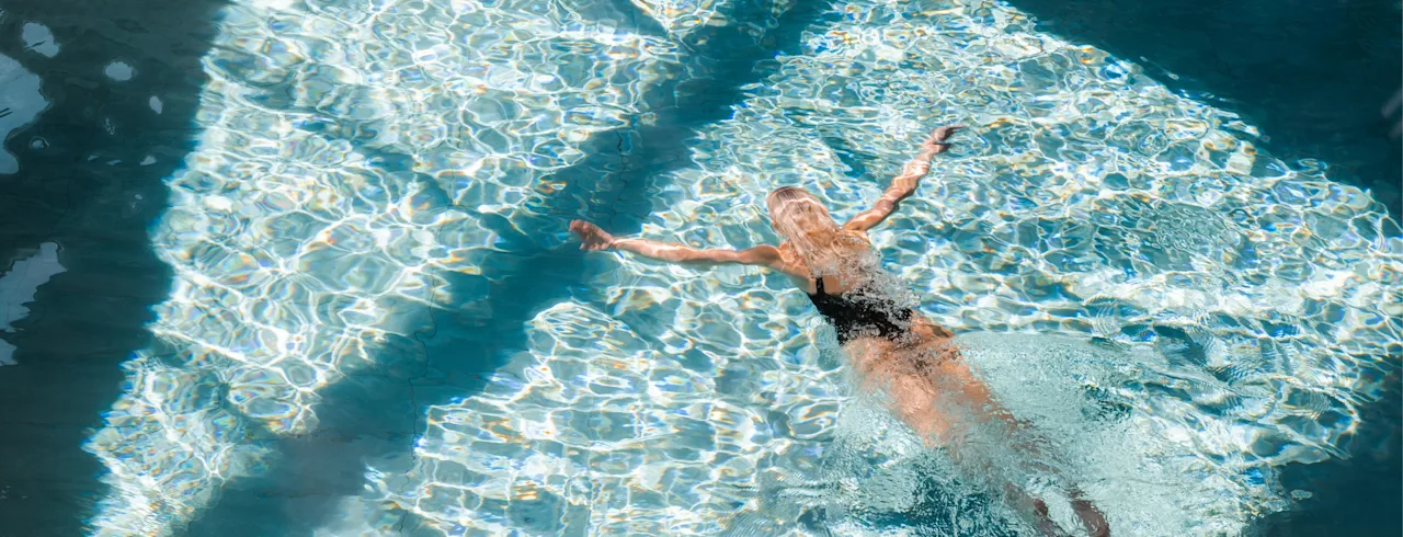 En person svømmer rygcrawl i en solbeskinnet pool og skaber krusninger og reflektioner i det klare vand.