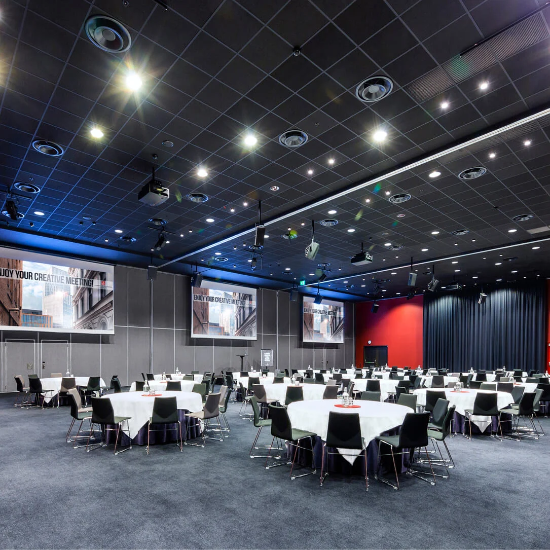 Runde borde med stoler er satt opp for et møte i et stort konferanserom med mørke gardiner og mønstret teppe. Banneret lyder "ENJOY YOUR CREATIVE MEETING!".
