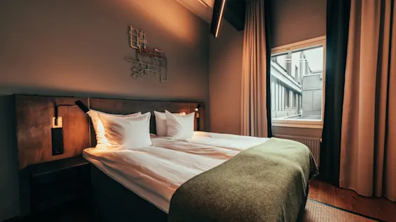 A cozy hotel room with a neatly made bed, warm lighting, and a window looking out onto a building.