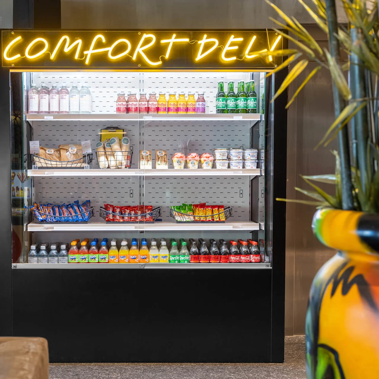 A refrigerated display case with snacks and drinks, labeled "COMFORT DELI!" in neon, is situated in a modern indoor space with decorative plants and umbrella stand.
